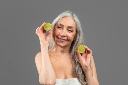 Foto de Smiling old caucasian lady with gray hair hold pieces of lemons, has fun, isolated on gray background, studio. Anti-aging treatment, health and skin care at home, moistening procedure and spa day - Imagen libre de derechos