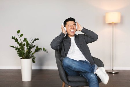 Photo for Cheerful emotional mature korean man in wireless headphones with open mouth sings song, enjoys music in living room interior with white wall. Relax and rest, technology for entertainment, fun at home - Royalty Free Image