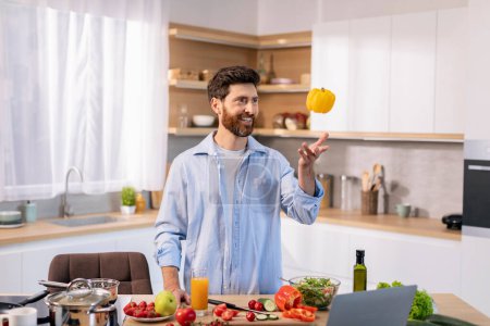 Photo for Cheerful millennial caucasian guy cooks eat, catches pepper, has fun at table with vegetables and computer in kitchen interior. Shooting video of new recipe for food blog and prepare meal at home - Royalty Free Image