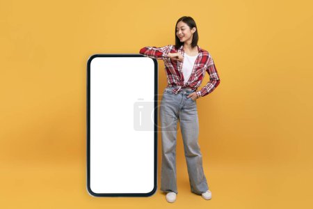 Téléchargez les photos : Happy japanese lady leaning on big smartphone with blank white screen and smiling, yellow studio background, mockup. Template for mobile app, website, your advertisement design - en image libre de droit