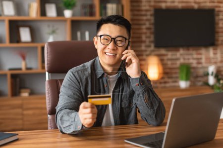 Foto de Smiling mature korean male in glasses calls by phone, uses credit card at workplace with laptop in office interior. Conversation with client, money management, checking finances accounts and shopping - Imagen libre de derechos
