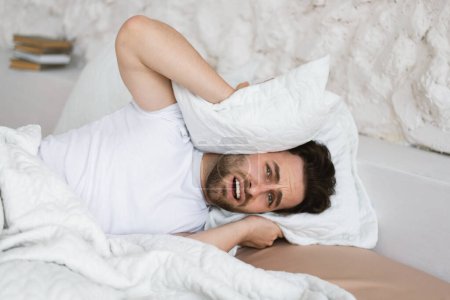 Photo for Tired angry sad young caucasian guy lies on bed, covers his ears with pillow, suffers from noisy neighbors, stress and depression in bedroom interior. Disease, human emotions and insomnia at home - Royalty Free Image