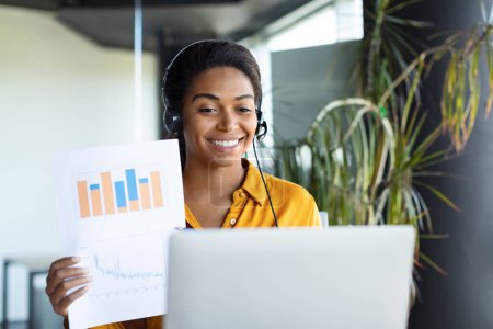 Photo for Happy black female entrepreneur sitting at table and showing paper with color charts in computer webcam in office interior. Designer work, business and modern tech, meeting and video call - Royalty Free Image