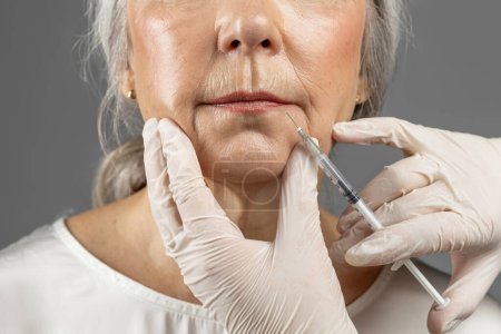 Foto de Serious mature european female on procedure, cosmetologist hands in gloves make botox injection, isolated on gray background, close up. Rejuvenation skin, professional treatment and anti-aging care - Imagen libre de derechos