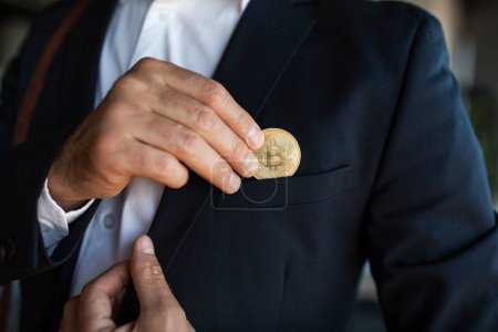 Téléchargez les photos : Male ceo manager in suit putting bitcoin coin in pocket, standing in office interior, closeup, cropped. Successful business, businessman recommending cryptocurrency, profit and earnings - en image libre de droit