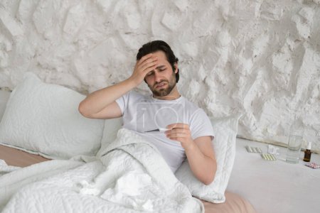 Téléchargez les photos : Unhappy sad young caucasian man suffers from headache and fever, look at thermometer, touch forehead with hand, sits on white bed in bedroom interior. Flu and cold, high temperature, health problems - en image libre de droit