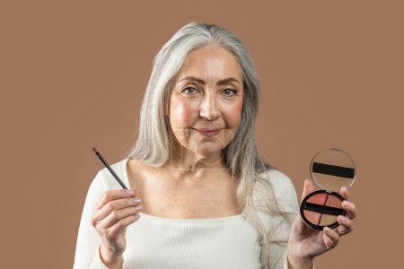 Téléchargez les photos : Cheerful senior european lady with gray hair shows cosmetics, blush or eye shadow with brush isolated on brown background, studio. Nude makeup lesson, daily procedures, beauty recommendation, advice - en image libre de droit