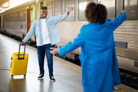 Foto de Black Couple Meeting On Platform At Railway Station After Train Arrival, Cheerful African American Man And Woman Happy To See Each Other, Guy Carrying Luggage And Spreading Hands For Hug - Imagen libre de derechos