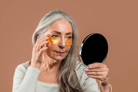 Téléchargez les photos : Serious elderly caucasian woman with natural beauty applies patches under her eyes and looks in mirror, isolated on brown background, studio. Beauty care, anti-aging procedures, fight against old skin - en image libre de droit