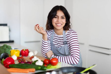 Téléchargez les photos : Positive cheerful attractive arabic young woman wearing grey apron chef posing at cozy kitchen at home, lady cooking healthy delicious food, using only eco organic vegetables, copy space - en image libre de droit