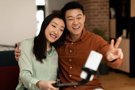 Téléchargez les photos : Excited korean couple middle aged man and young woman taking selfie with selfie stick, while spending time together at home. Spouses smiling at cellphone webamera - en image libre de droit