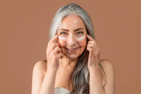 Téléchargez les photos : Glad elderly caucasian female with natural beauty applies cream on her face, fight with wrinkles, enjoy anti-aging procedure, isolated on brown background, close up, studio. Beauty care, spa treatment - en image libre de droit
