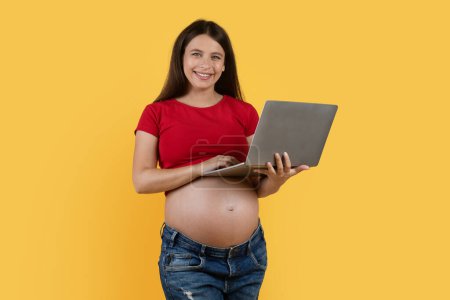 Téléchargez les photos : Smiling Pregnant Female Using Laptop Computer For Online Shopping While Standing Isolated Over Yellow Background, Cheerful Young Expectant Woman Enjoying Purchasing In Internet, Copy Space - en image libre de droit