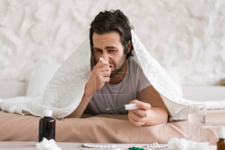 Photo for Despaired sad young caucasian sick male lies on bed under blanket, suffers from runny, blows his nose in napkin in white bedroom interior. Illness, flu and cold, bad feeling and treatment at home - Royalty Free Image