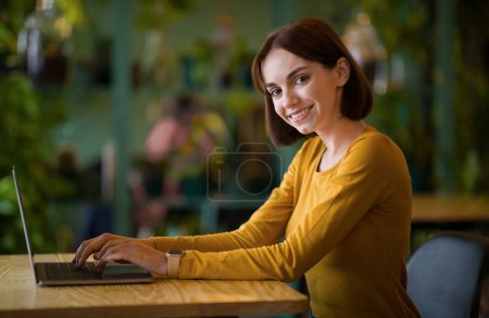 Téléchargez les photos : Women in business. Side view of confident cheerful young brunette woman in smart casual wear freelancer working on modern laptop while sitting at table in creative office or cafe, copy space - en image libre de droit