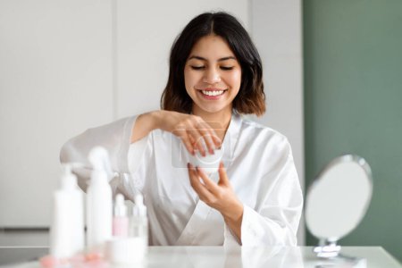 Foto de Cheerful happy pretty brunette young middle eastern woman in white silk bathrobe using face cream at home, beautiful lady testing newest organic beauty product, sitting at vanity table, copy space - Imagen libre de derechos
