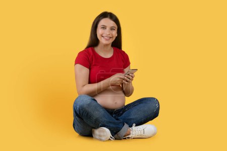 Foto de Nice App. Portrait Of Beautiful Young Pregnant Woman With Smartphone In Hands Sitting On Floor Over Yellow Background, Smiling Expectant Female Using Mobile Phone For Shopping Or Communication - Imagen libre de derechos