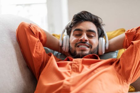 Photo for Closeup candid shot of joyful attractive bearded middle eastern young man in orange shirt lying on couch with hands behind head, listening to music with closed eyes and smiling, home interior - Royalty Free Image