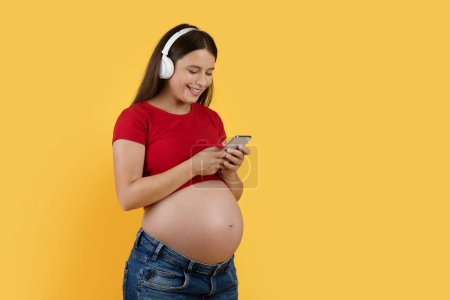Foto de Modern Technologies. Happy Pregnant Woman In Wireless Headphones Listening Music Via App On Smartphone And Messaging With Friends, Smiling Expectant Lady Standing Over Yellow Background, Copy Space - Imagen libre de derechos