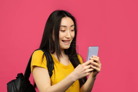 Photo for Happy asian female student using smartphone, texting online while standing with backpack over pink background, copy space. Woman using educational mobile app on cellphone - Royalty Free Image