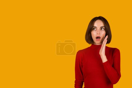 Foto de Amazed brunette young caucasian woman with cute hairstyle in red looking at copy space for advertisement and touching her face over orange studio background, web-banner. Nice offer, discount, news - Imagen libre de derechos