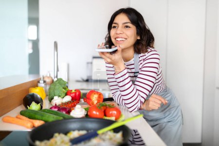 Téléchargez les photos : Happy pretty short-haired brunette young middle eastern woman recording voice message using smartphone while cooking healthy organic food at home, looking at copy space and smiling, kitchen interior - en image libre de droit