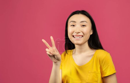 Téléchargez les photos : Happy japanese lady gesturing victory symbol and smiling to camera, standing on pink studio background, free space. Portrait of asian female showing v-sign, peace gesture - en image libre de droit