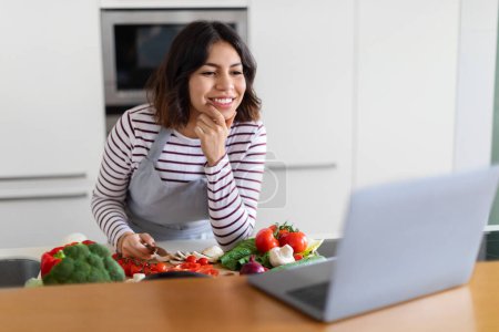 Photo for Excited beautiful brunette young middle eastern woman enjoying cooking at home, reading recipe online, checking food blog on Internet, using laptop while preparing healthy meal at kitchen, copy space - Royalty Free Image