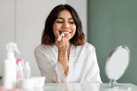 Foto de Pretty young middle eastern woman looking at mirror, using lipstick, home interior, empty space. Attractive lady taking care of her beautiful lips, putting nourishing balm, applying makeup, headshot - Imagen libre de derechos