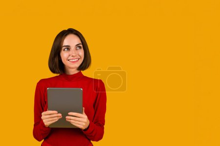 Foto de Nice online offer, deal. Cheerful pretty young brunette woman in red checking newest mobile application, using modern digital tablet and looking at copy space over orange studio background, smiling - Imagen libre de derechos