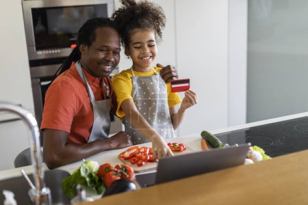Téléchargez les photos : Happy Black Father And Daughter Using Laptop And Credit Card While Cooking Lunch In Kitchen, Smiling African American Dad And Female Child Ordering Groceries Online, Making Internet Purchases At Home - en image libre de droit