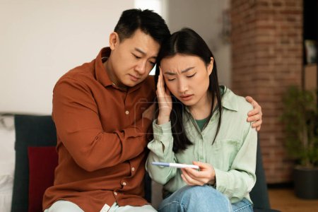 Photo for Upset mature asian husband comforting his depressed young wife with negative pregnancy test, sitting on sofa at home. Korean couple cannot have baby, suffering from infertility indoors - Royalty Free Image