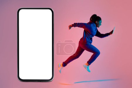Téléchargez les photos : Active black lady exercising in the morning, running with cellphone next to giant smartphone with blank white screen on pink neon background, mockup without copy space - en image libre de droit