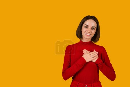 Foto de Thankful pretty young woman in red holding hands over chest and smiling, showing love and loyalty over orange studio background, sharing feelings, expressing gratitude, copy space for advert - Imagen libre de derechos