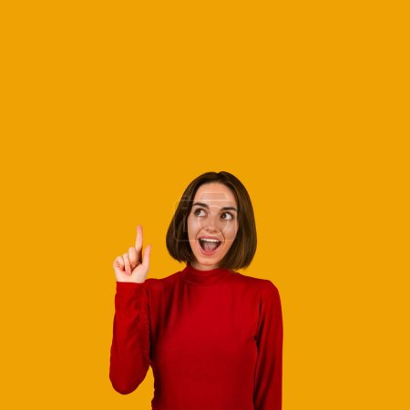 Téléchargez les photos : Happy pretty brunette young woman in red pointing upwards at copy space showing idea or eureka gesture and smiling, generating interesting plan. Studio shot over orange background - en image libre de droit