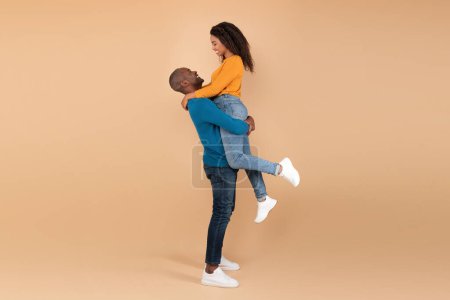 Téléchargez les photos : Loving mature black man lifting his wife and holding her on peach studio background, profile side view, free space. Romantic adult couple expressing their love to each other - en image libre de droit