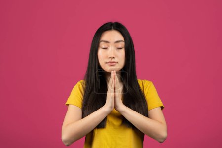Foto de Pray and meditate. Portrait of chinese lady keeping palms together and praying, meditating, having calm and peaceful facial expression, pink background, studio shot - Imagen libre de derechos