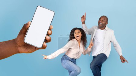 Foto de Great offer. Happy black couple dancing and looking at giant cell phone showing blank space for mock up. Hand holding white empty screen, blue studio background wall - Imagen libre de derechos