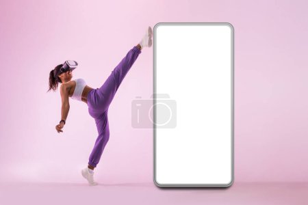 Photo for Sporty young black lady in vr googles exploring virtual reality fitness game near huge smartphone with free space for mockup. Energetic woman throwing kick - Royalty Free Image