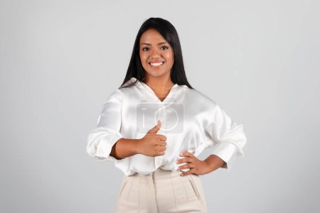 Foto de Happy confident millennial black woman in white blouse show thumb up, isolated on gray background, studio, copy space. Agreement, great deal business, work, study and recommendation from professional - Imagen libre de derechos