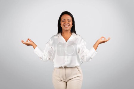 Téléchargez les photos : Glad serene millennial black female in white blouse with closed eyes meditation, enjoy spare time, isolated on gray background, studio. Recommends rest, relax, break in business, work and startup - en image libre de droit