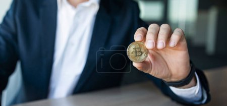 Téléchargez les photos : Closeup shot of businessman showing bitcoin, sitting at workplace in office, selective focus on golden coin, panorama with copy space. Cryptocurrency concept - en image libre de droit