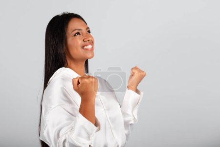 Photo for Cheerful millennial black businesswoman in white blouse make success, victory gesture, looking at free space, isolated on gray background, studio. Winning, great deal, business decision, ad and offer - Royalty Free Image