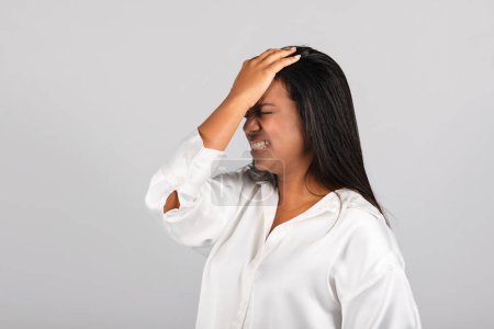 Photo for Despaired frustrated millennial black female in white blouse presses hand to forehead, suffering from stress, headache, mistake, isolated on gray background. Problems at business, work and deadline - Royalty Free Image