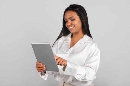 Photo for Cheerful confident millennial black businesswoman in white blouse typing on tablet isolated on gray background, studio. Device for business, app for work, study, investment and startup, ad and offer - Royalty Free Image