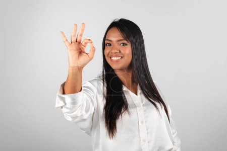 Foto de Cheerful millennial black lady in white blouse show ok sign with hand, isolated on gray background, studio. Great deal, business, work, study, recommendation from professional, advice and agreement - Imagen libre de derechos