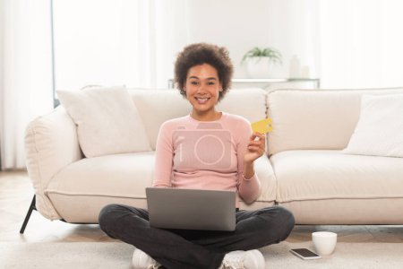 Téléchargez les photos : Cheerful young mixed race curly female shopaholic sit on floor with laptop, showing credit card, enjoy huge sale in living room interior. Work, business, finance, banking and online shopping at home - en image libre de droit