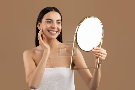 Téléchargez les photos : Beautiful half-naked young woman with long brunette hair wrapped in bath towel looking at mirror, pretty smiling lady touching her silky smooth face skin, isolated on beige studio background - en image libre de droit
