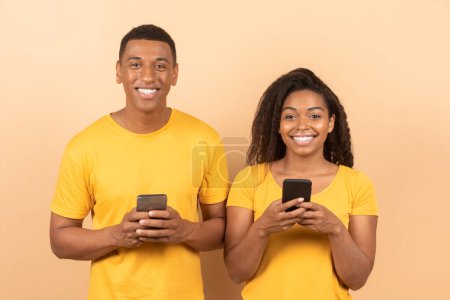 Téléchargez les photos : Portrait of happy black couple using cellphones and smiling at camera, standing on yellow studio background. African american spouses using new app and browsing internet - en image libre de droit