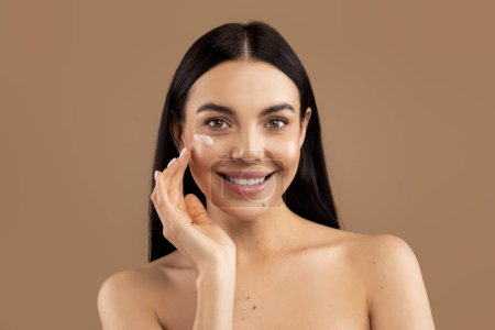 Foto de Cheerful sensual naked long-haired brunette millennial woman with smile on her face using facial eye cream for young smooth glowing skin, isolated on beige studio background, closeup shot - Imagen libre de derechos